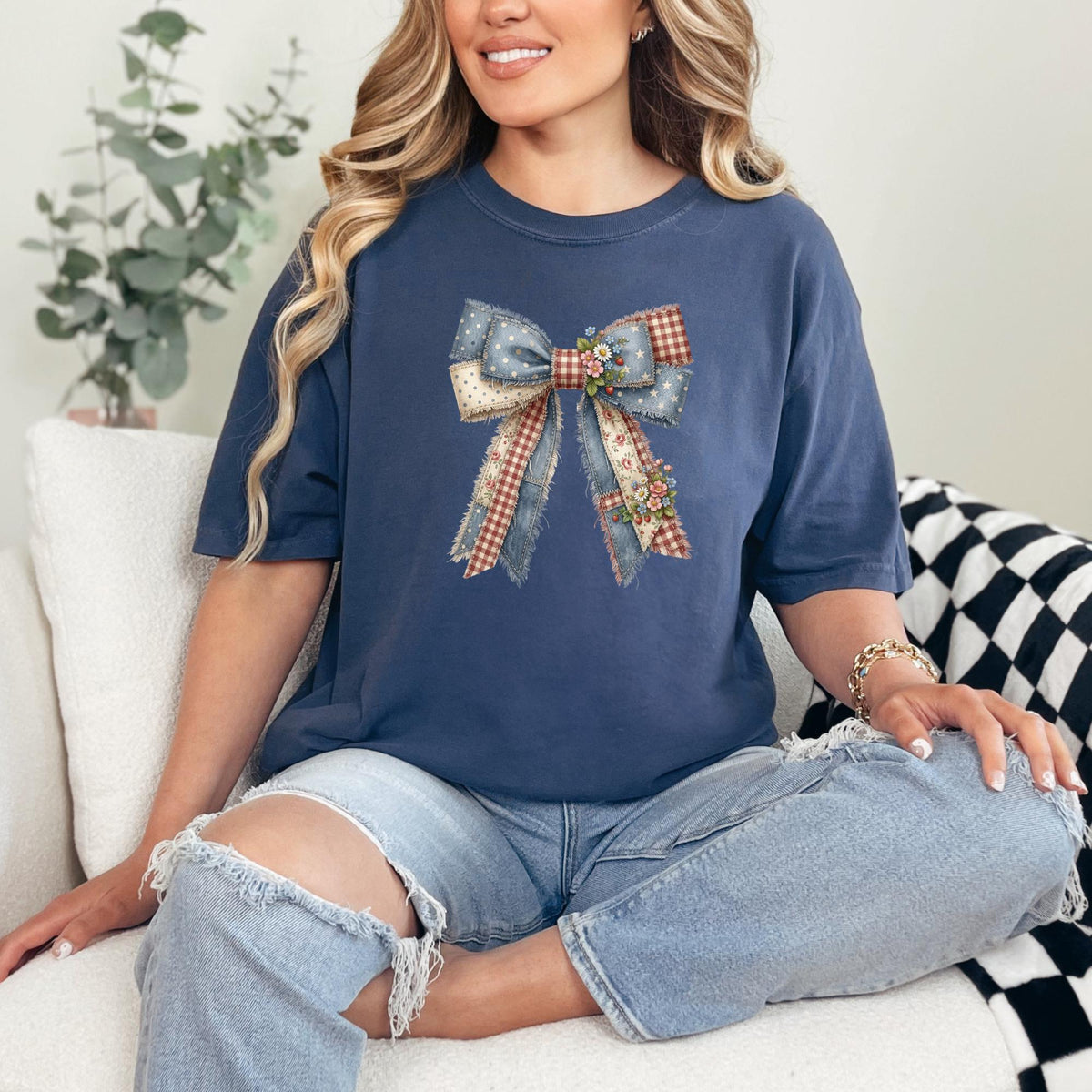 A woman sitting on a white couch wearing a navy blue t-shirt with a large denim and gingham bow graphic on the front, paired with distressed light-wash jeans.