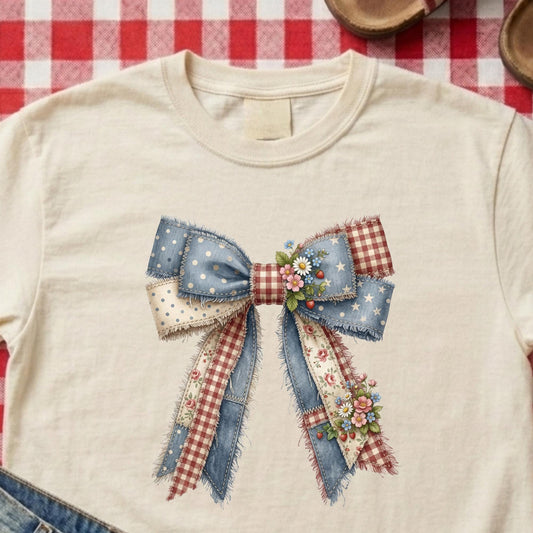 A cream-colored short-sleeve t-shirt featuring a large, detailed patchwork bow graphic on the chest. The bow is made from various fabrics including blue denim with white stars, red gingham, polka-dot cream ribbon, and small floral clusters.
