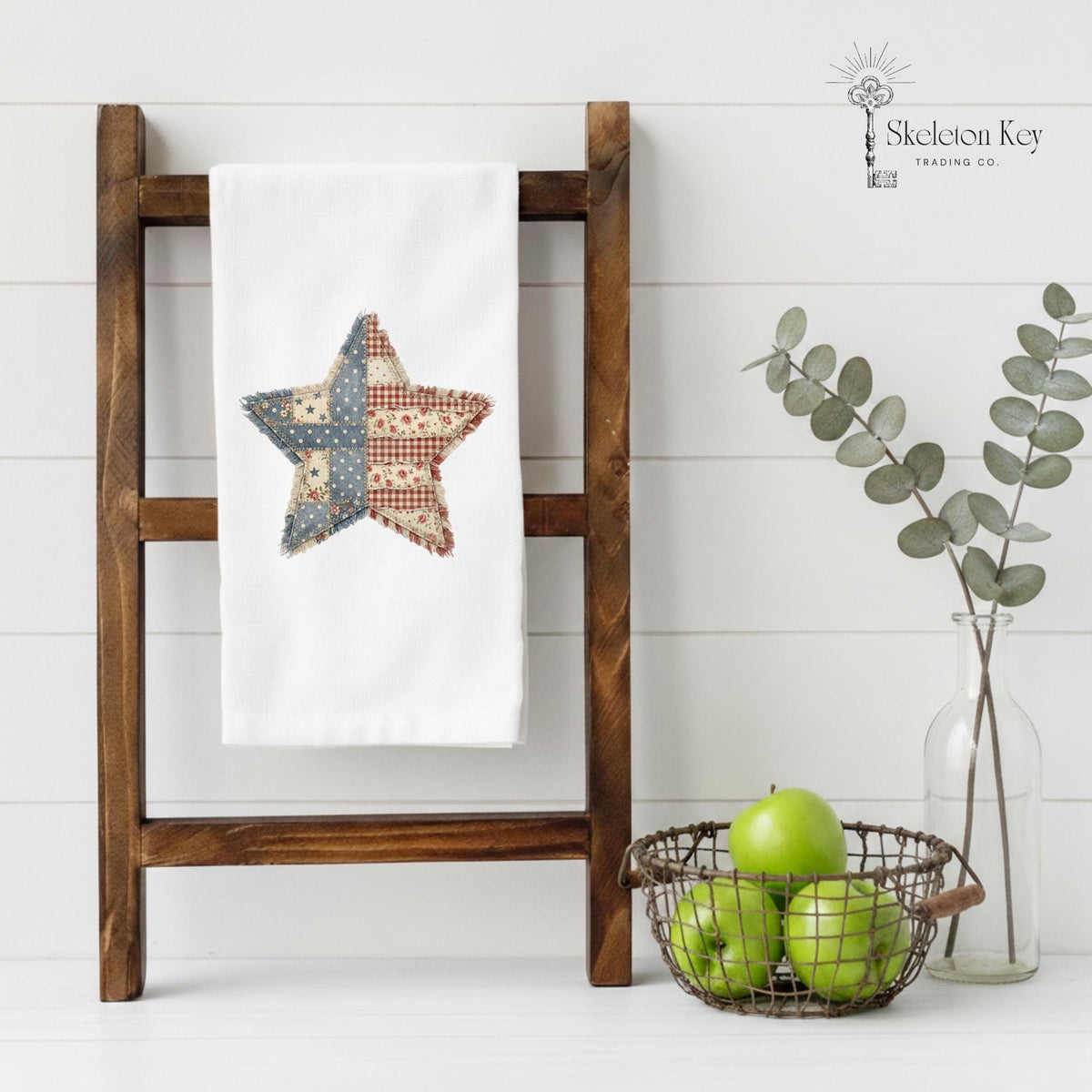 A white kitchen towel with a rustic patchwork star design in red, blue, and beige, hanging on a dark wooden ladder shelf. The scene is styled with a wire basket of green apples and a glass vase with eucalyptus branches against a white shiplap wall.