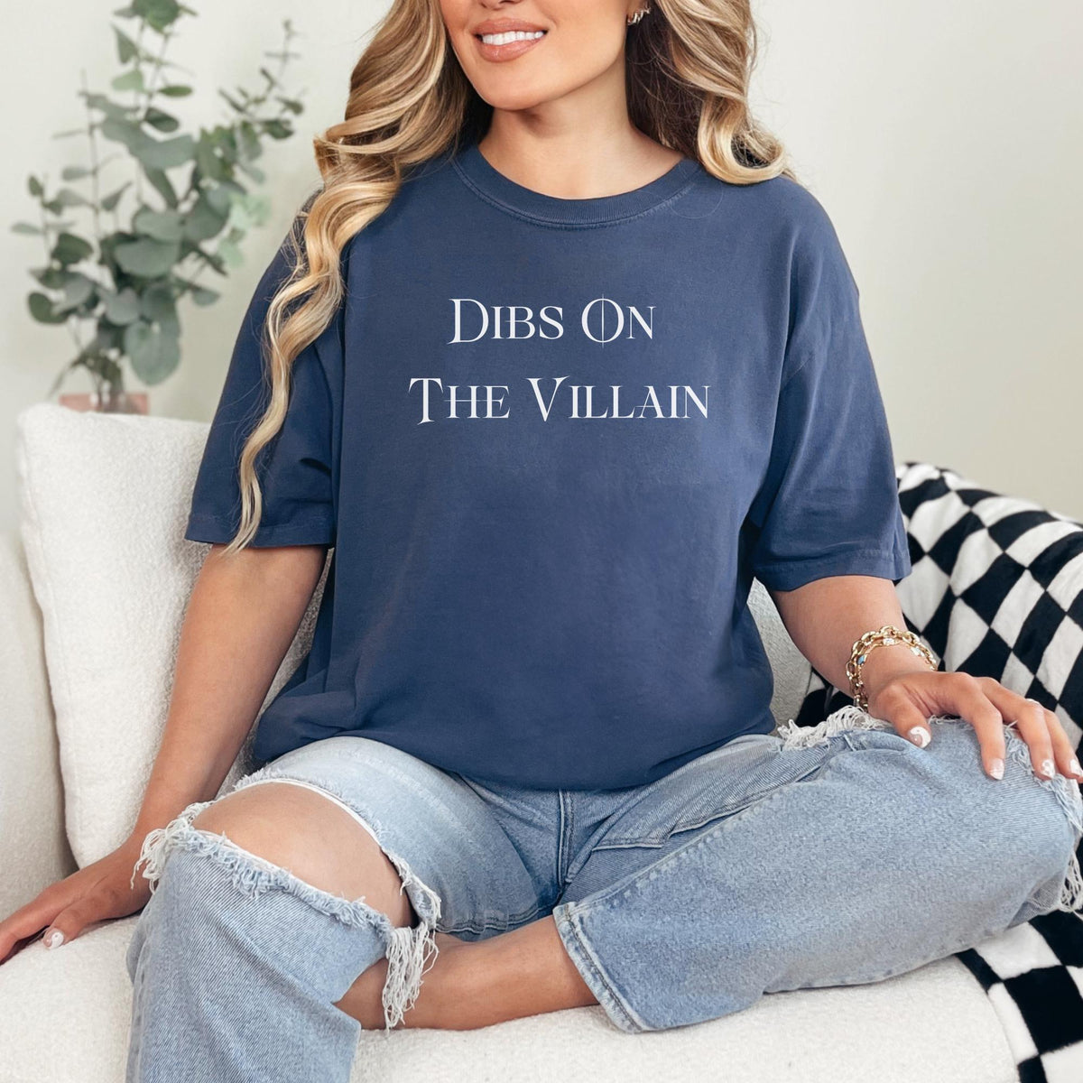 A woman sitting on a white couch wearing a navy blue graphic t-shirt with the text 'DIBS ON THE VILLAIN' in white serif font, paired with light-wash ripped jeans.