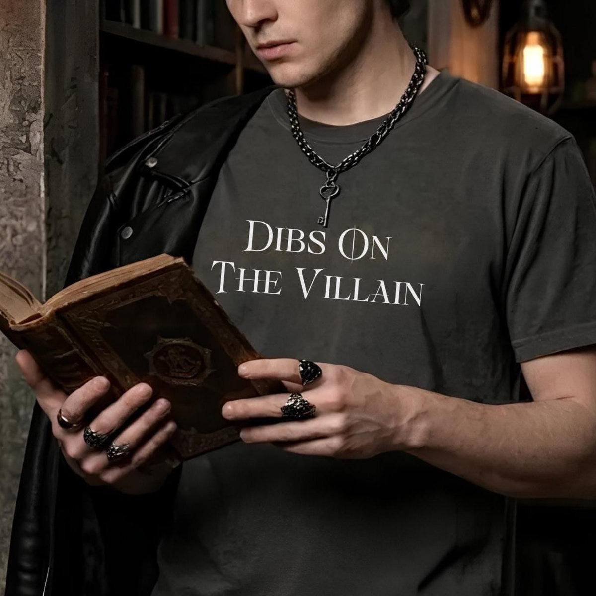 A man wearing a dark gray graphic t-shirt with the text 'DIBS ON THE VILLAIN' in white, styled with a black leather jacket, silver chain necklace with a key pendant, and multiple rings. He is holding an open antique book in a dimly lit, atmospheric library setting.