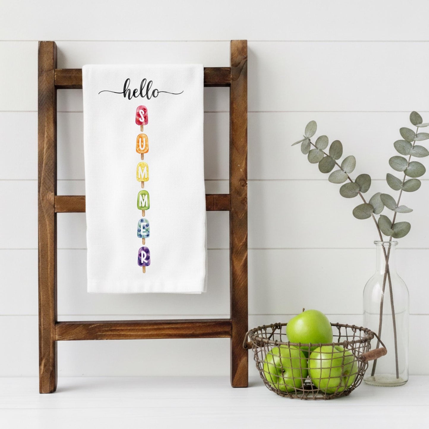White towel with colorful letters spelling 'hello summer' on a wooden ladder, with a basket of apples and a vase with eucalyptus leaves in the background.
