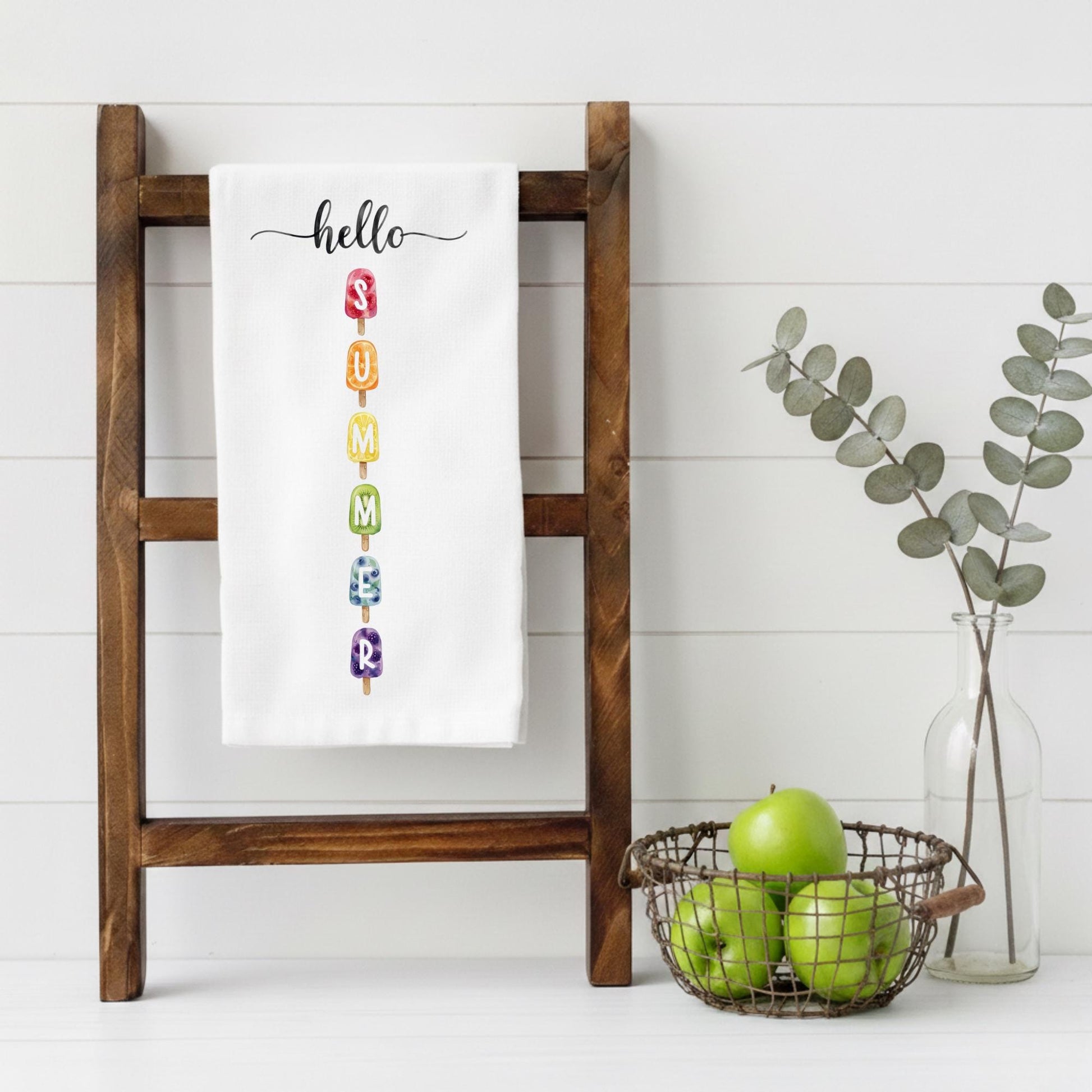 White towel with colorful letters spelling 'hello summer' on a wooden ladder, with a basket of apples and a vase with eucalyptus leaves in the background.