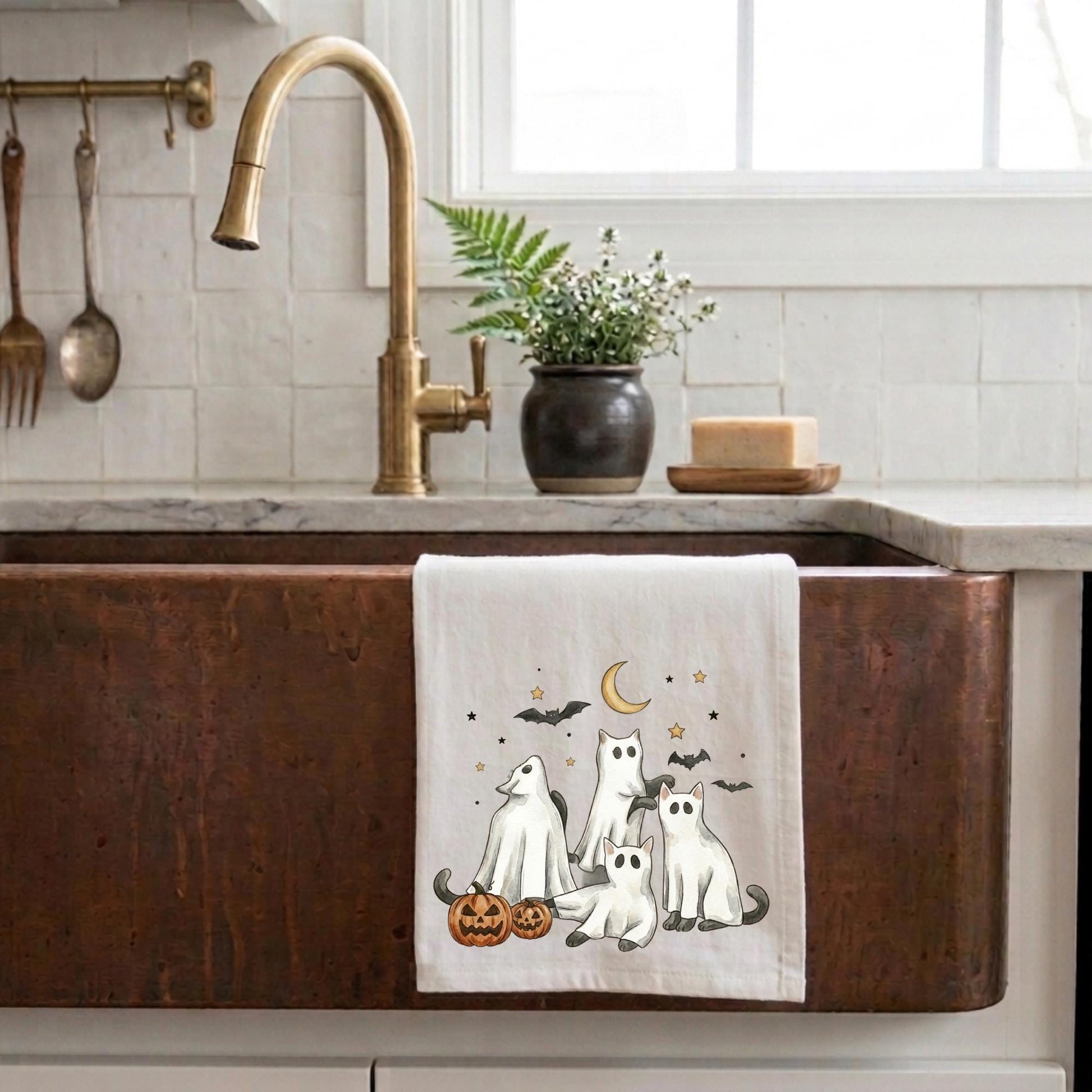 Cute ghost cat Halloween tea towel with four ghost cats, jack-o'-lanterns, bats, and crescent moon illustration hanging over a kitchen sink