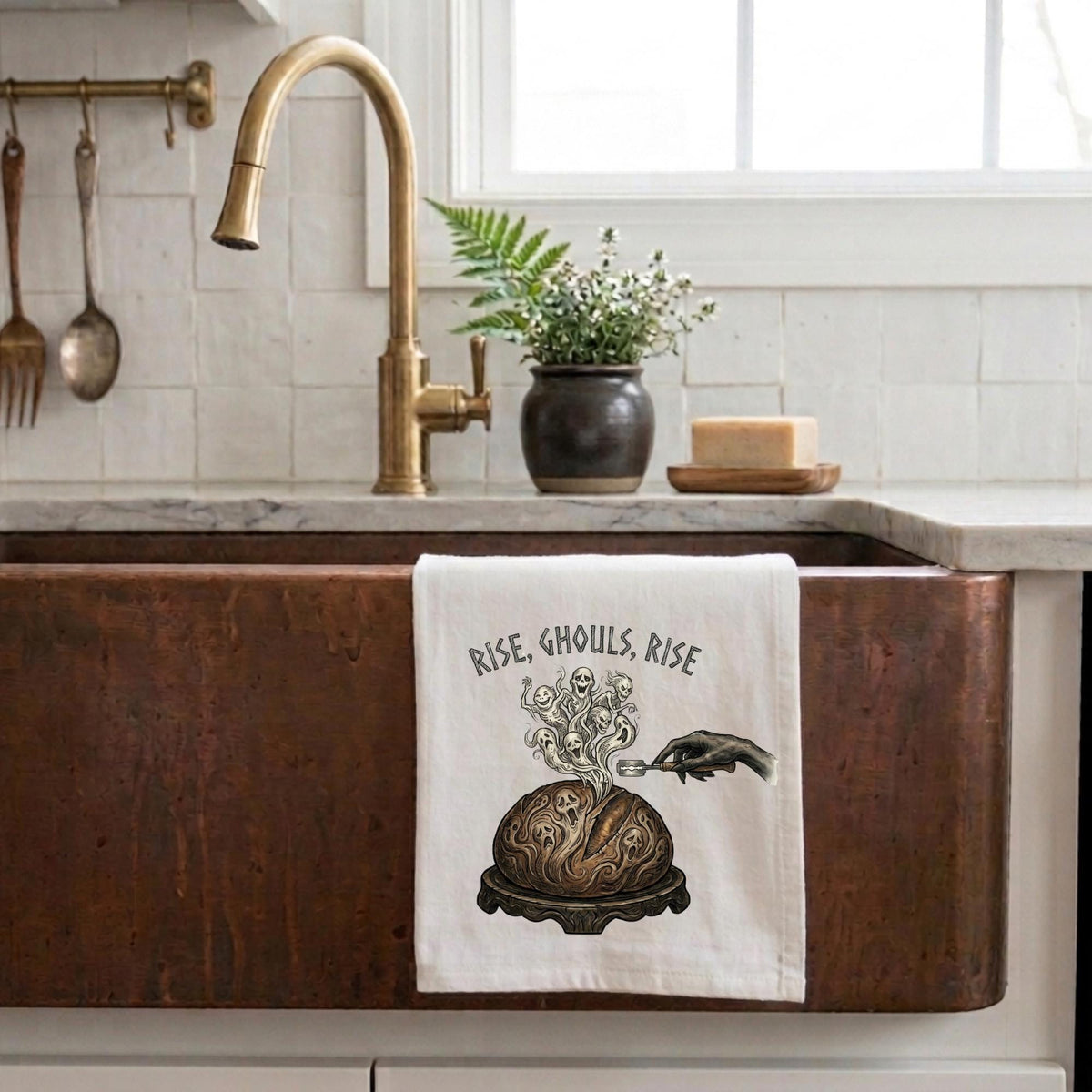 A white kitchen towel with a black graphic of a smoking cauldron and the text 'RISE, GHOULS, RISE' hanging over a copper farmhouse sink in a bright kitchen.