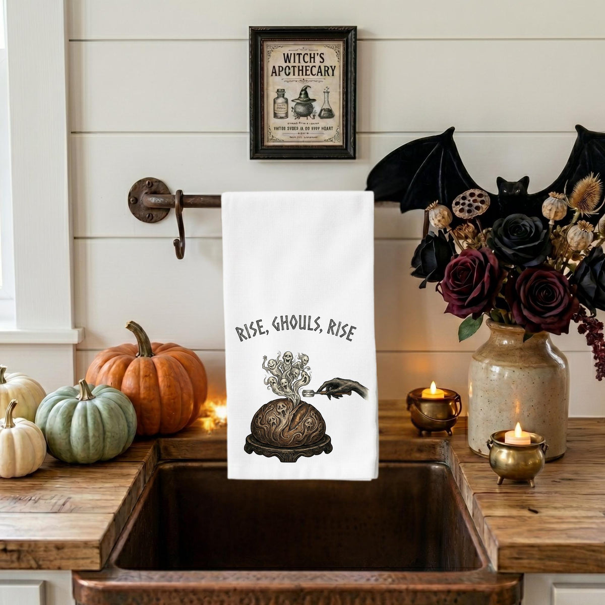 A white kitchen towel with a black gothic illustration and the text 'RISE, Ghouls, RISE' hangs over a rustic copper sink. The scene is styled for Halloween with pumpkins, a black bat decoration, a vase of dark roses, and candles on a white shiplap wall.