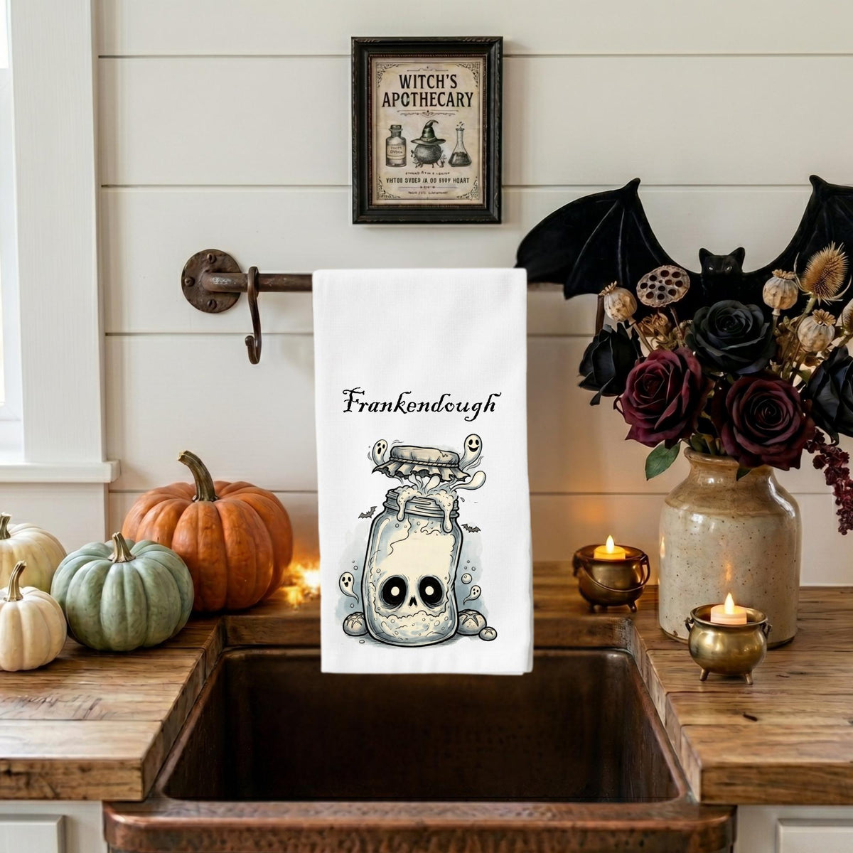 A white kitchen towel with a Halloween-themed illustration of a jar containing a skull and a small creature, printed with the name 'Frankendough'. The towel is hanging on a rustic metal rack above a dark copper sink in a decorated kitchen setting with pumpkins, candles, and a vase of dark roses.