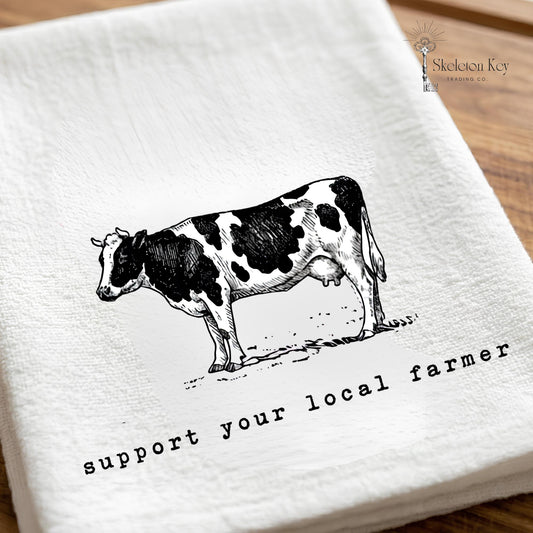 May include: White kitchen towel with a black and white illustration of a cow. The text "support your local farmer" is printed below the cow. The towel is made of a soft, textured fabric.