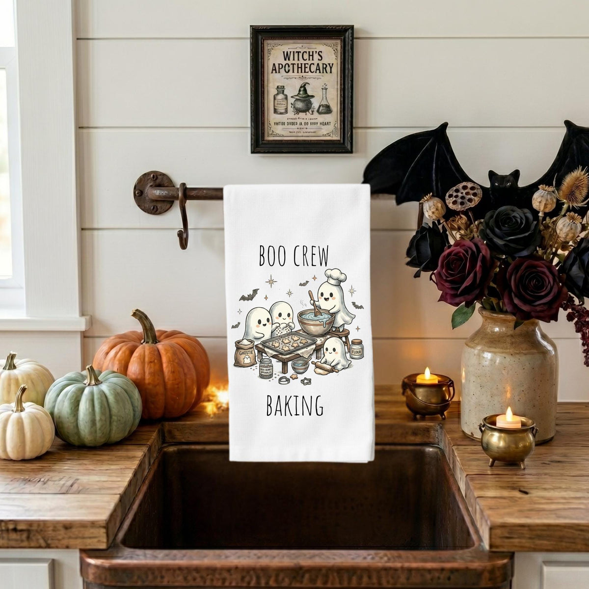 A white kitchen towel with a Halloween baking illustration and the words 'BOO CREW BAKING' hangs over a rustic sink in a styled kitchen scene with pumpkins, candles, a vase of dark flowers, and a framed 'Witch's Apothecary' sign on the wall.