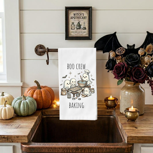 A white kitchen towel with a Halloween baking illustration and the words 'BOO CREW BAKING' hangs over a rustic sink in a styled kitchen scene with pumpkins, candles, a vase of dark flowers, and a framed 'Witch's Apothecary' sign on the wall.