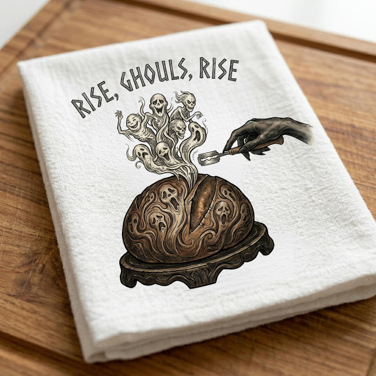 A folded white kitchen towel with a gothic illustration of a smoking brain, skulls, and the text 'RISE, GHOULS, RISE', placed on a wooden surface.