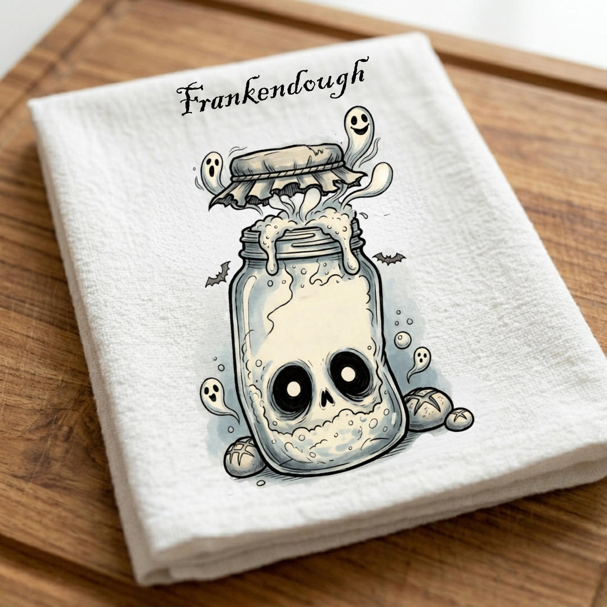 A folded white kitchen towel with a Halloween-themed graphic of a skull in a mason jar, ghosts, bats, and the word 'Frankendough' printed above, displayed on a wooden surface.