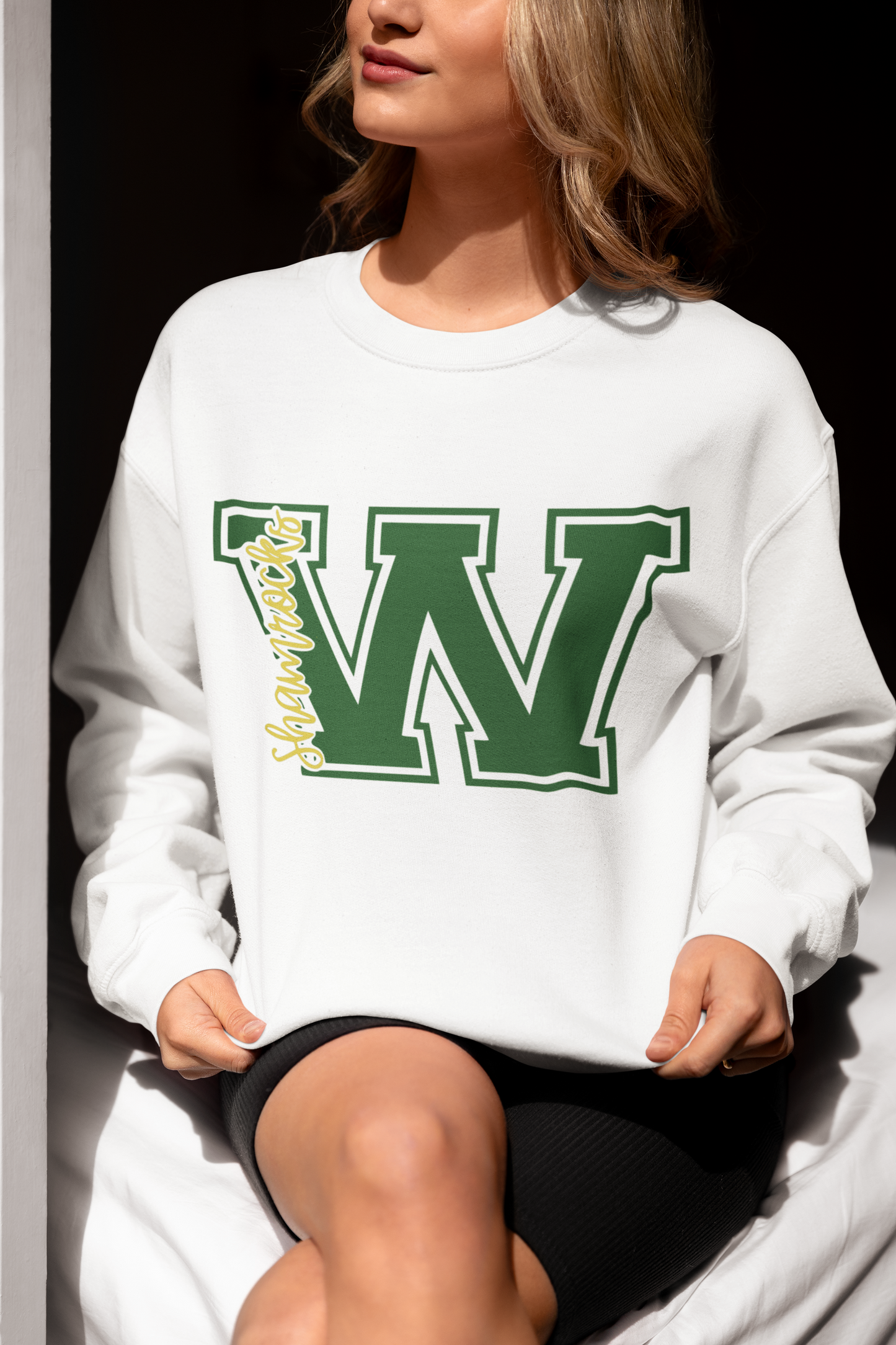 Woman sitting in soft natural light wearing a white Westfield Shamrocks varsity ‘W’ crewneck sweatshirt with green and yellow lettering.
